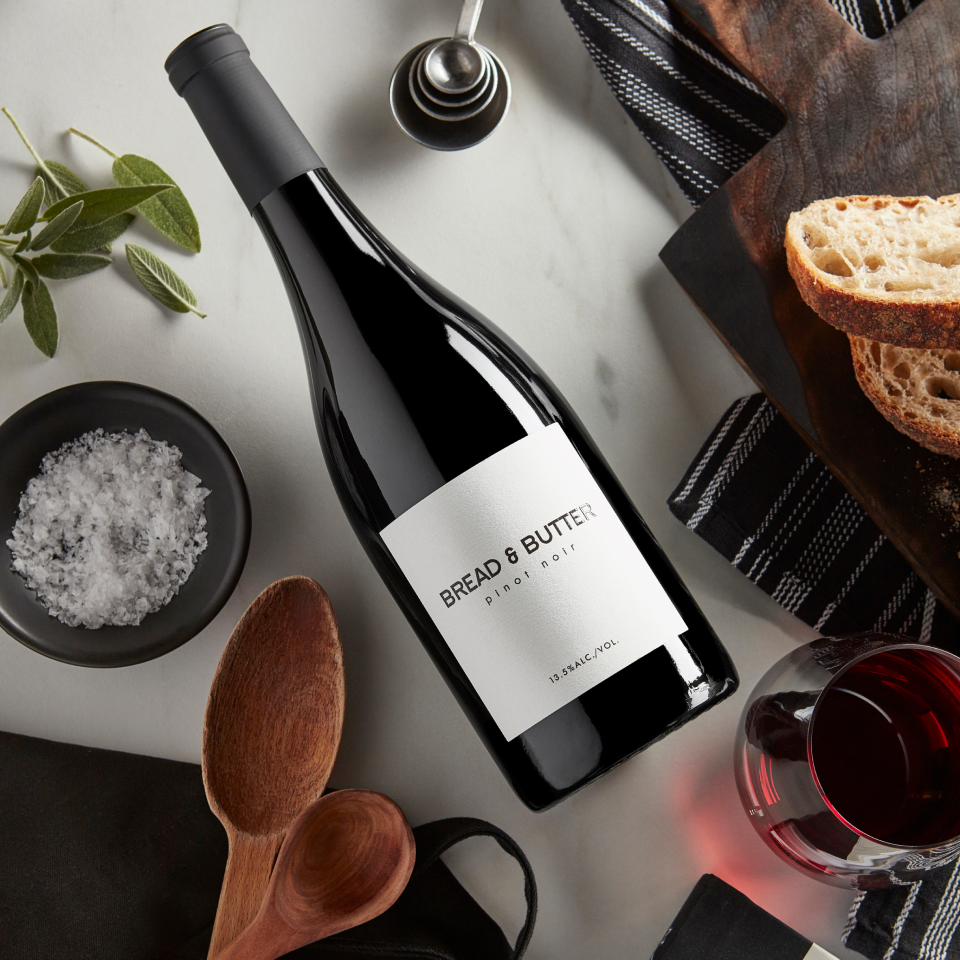 Bread & Butter Pinot Noir on a table with sage, salt, cooking tools, bread and a glass of wine poured.
