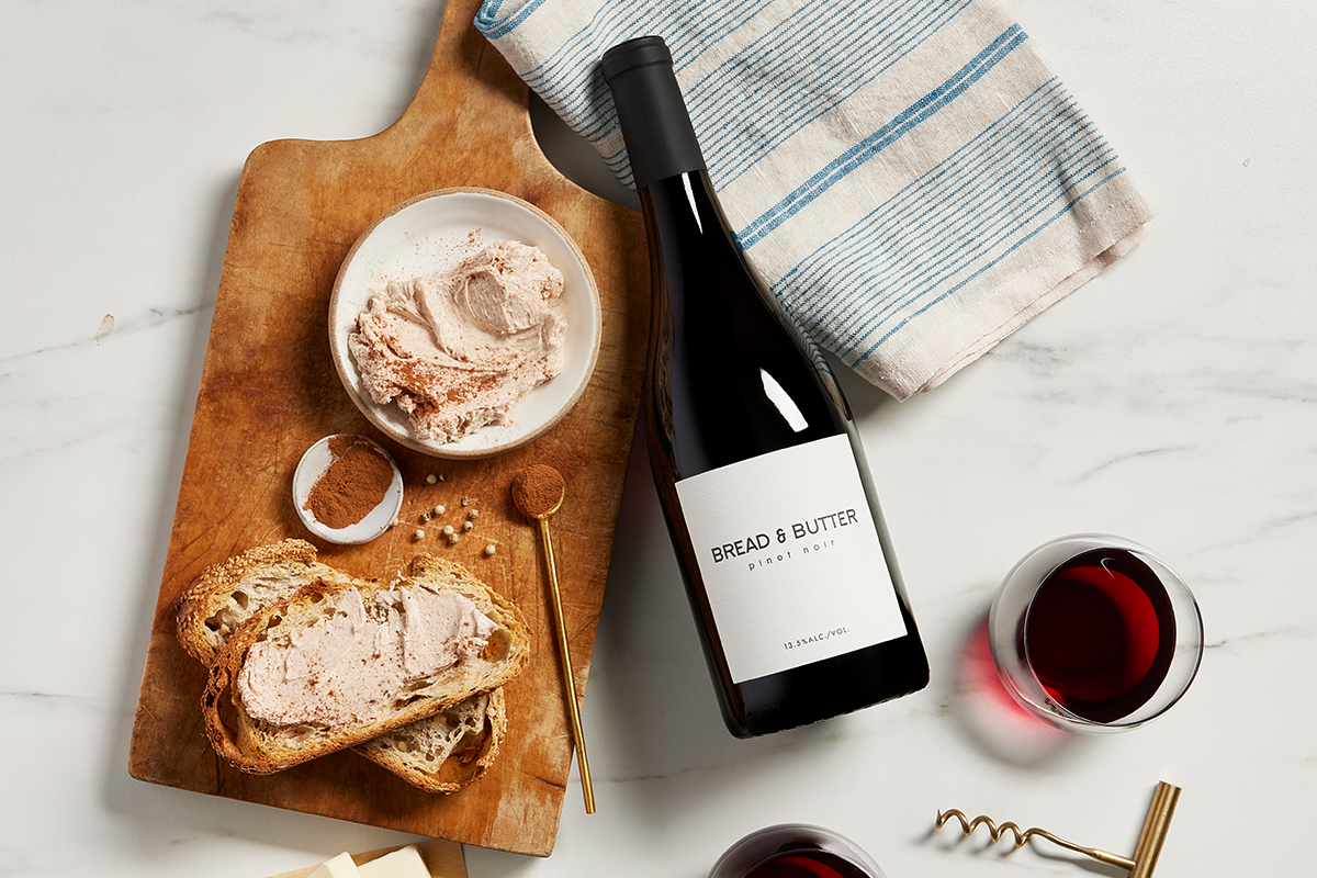 Bread & Butter Pinot Noir on a marble table with a bowl of Cinnamon Sugar Butter, sliced bread and a decorative spoon of cinnamon on a wood cutting board.
