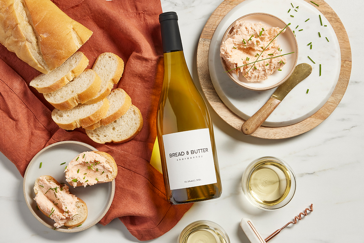 Bread & Butter Chardonnay on a marble background with a terracotta linen napkin next to a bowl of smoked salmon butter with slices of French bread and glasses of wine poured.