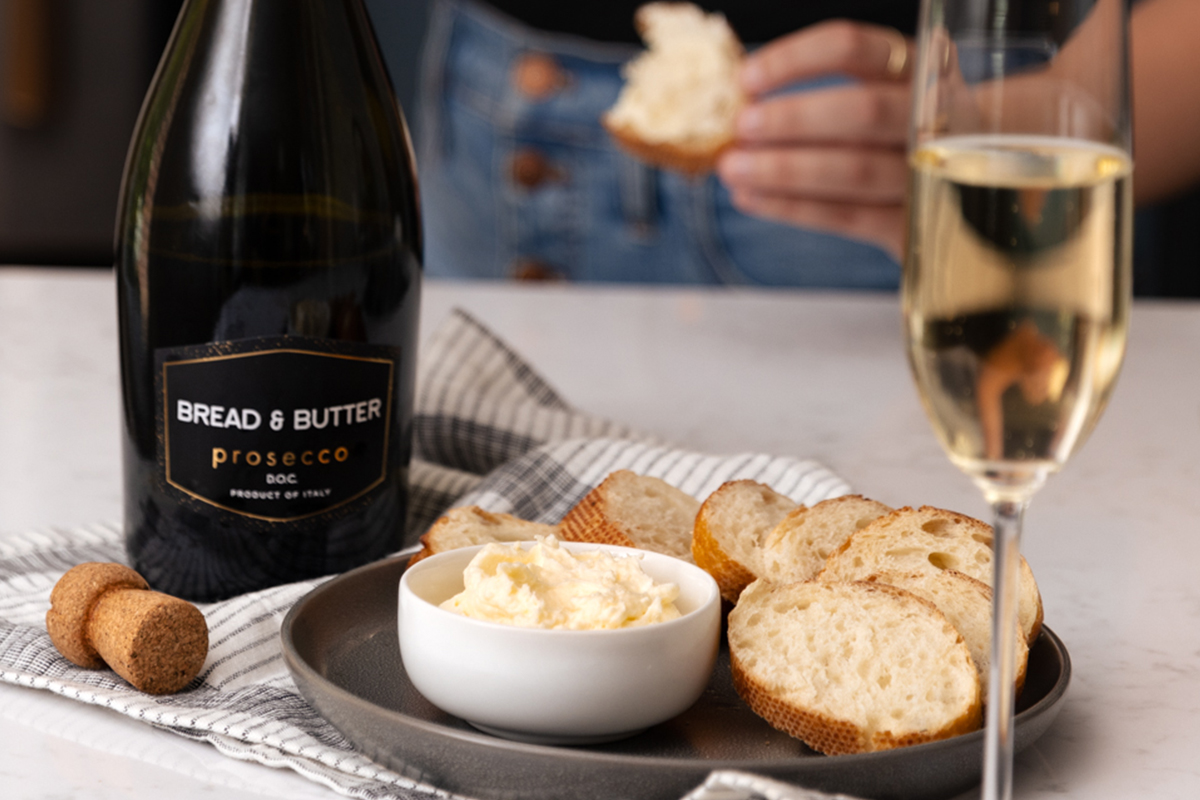 Bread & Butter Prosecco on a table with a napkin next to a bowl of Asiago Butter with a glass poured.
