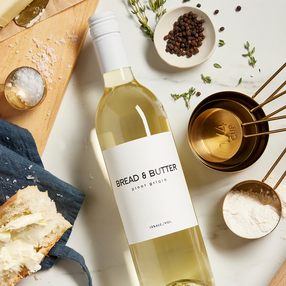 Bread & Butter California Pinot Grigio on a marble table with decorative brass measuring cyps, herbs, a bowl of peppercorns, flour, salt and a torn piece of bread with butter.