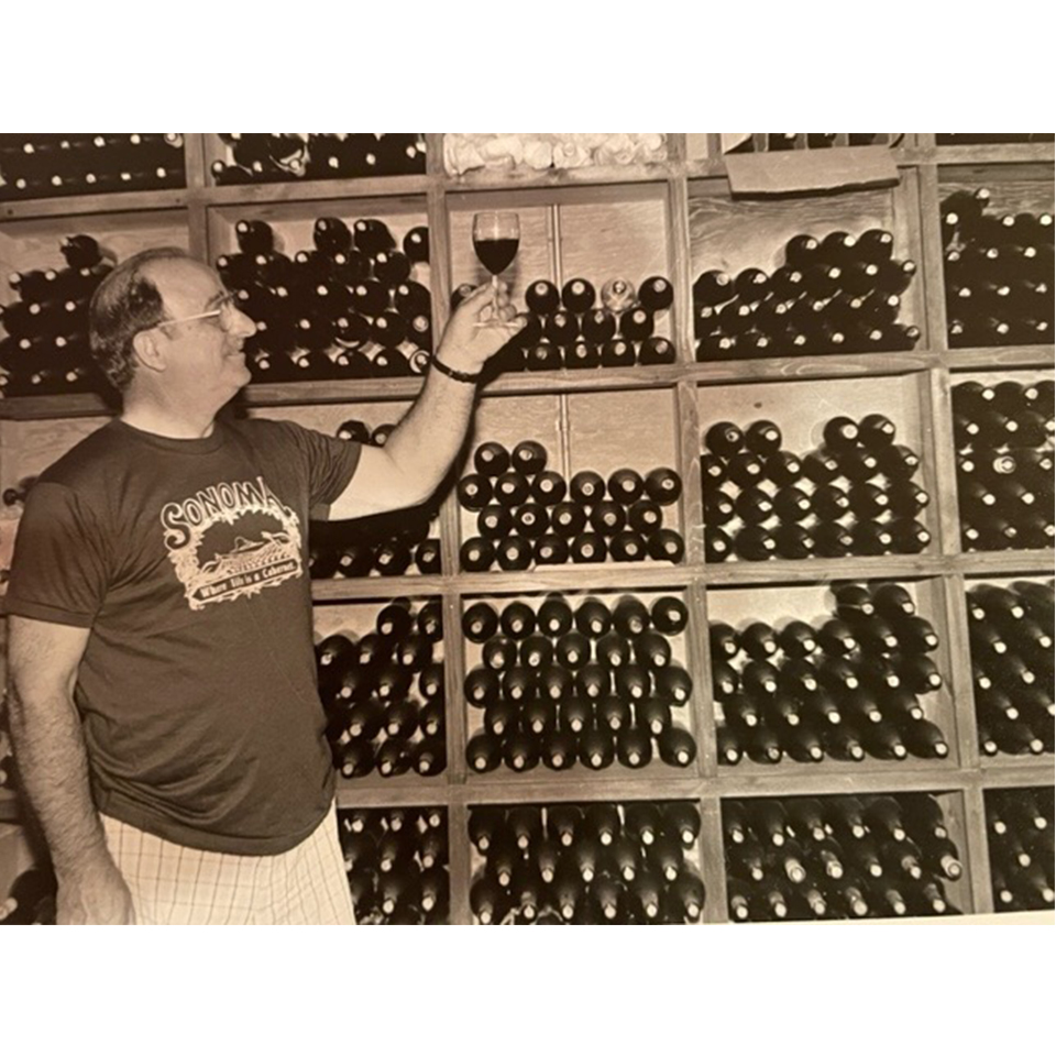 Linda Trotta's paternal grandfather, Frank, holding a glass of red wine in his basement winery/cellar filled with bottles of wine.